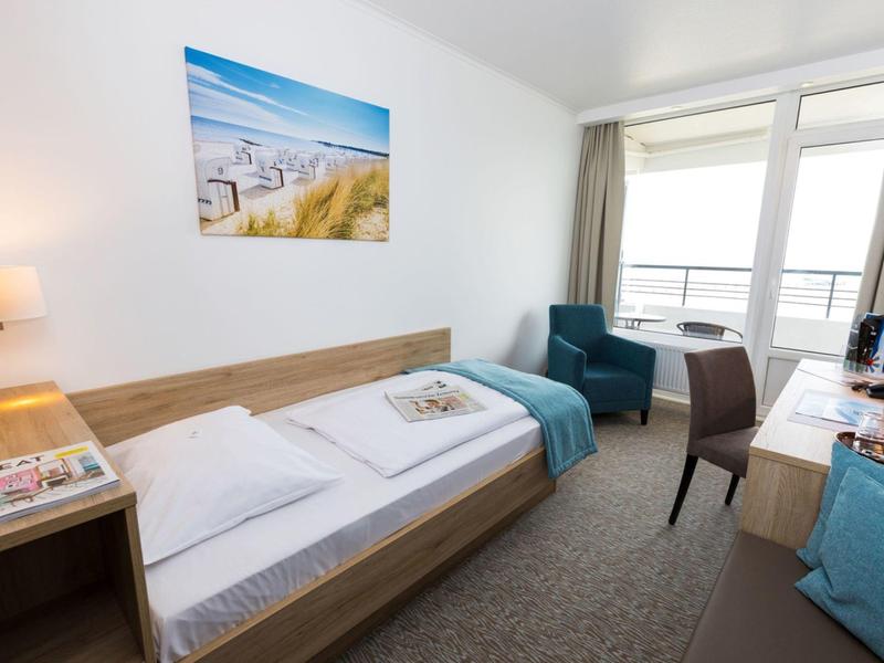 Helles Hotelzimmer mit Einzelbett, Sessel, Schreibtisch und Balkon mit Blick nach draußen.