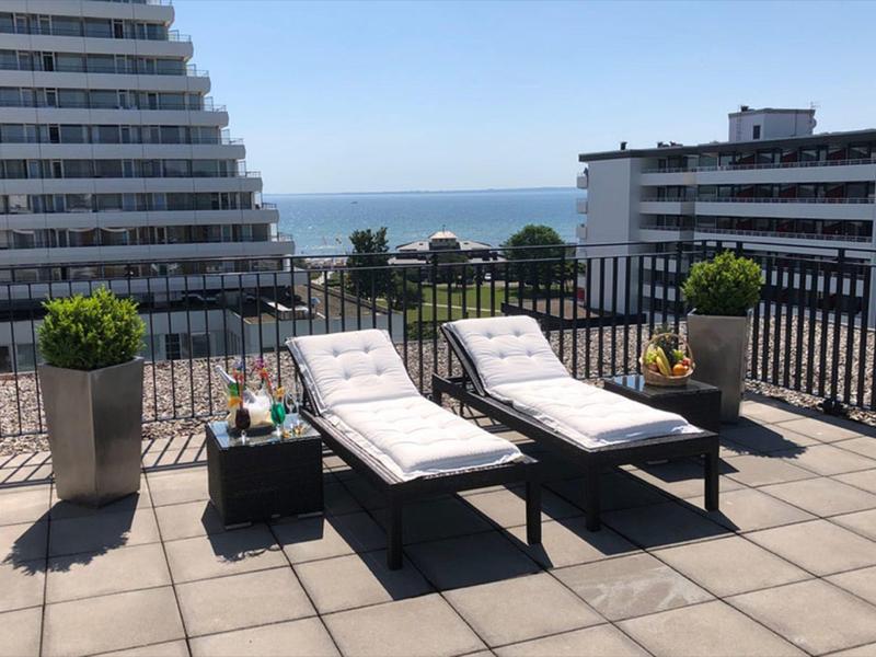 Zwei Liegestühle auf einer Dachterrasse mit Blick auf das Meer und umliegende Gebäude.