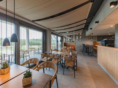 Moderní restaurace s velkými okny, dřevěnými stoly a židlemi a rostlinami na stolech.