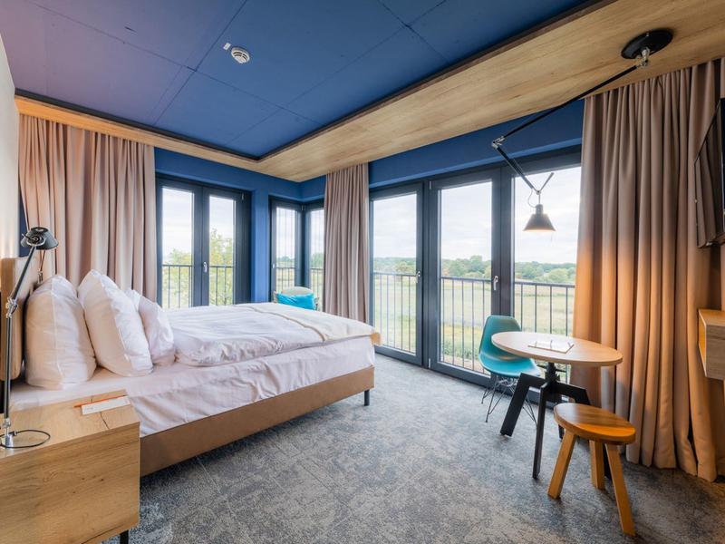 Chambre d'hôtel moderne avec lit double, grandes fenêtres et vue sur le paysage vert.