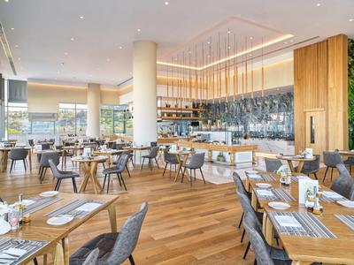 Modernes Restaurant mit Holzboden, weißen Säulen, grauen Stühlen und großen Fenstern.