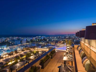 Stadtblick bei Dämmerung mit beleuchteten Straßen, Gebäuden und einer Terrasse mit Sitzgelegenheiten.