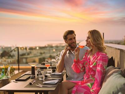 Paar sitzt draußen auf Terrasse, trinkt Wein, Blick auf Stadt im Sonnenuntergang, entspannte Atmosphäre.