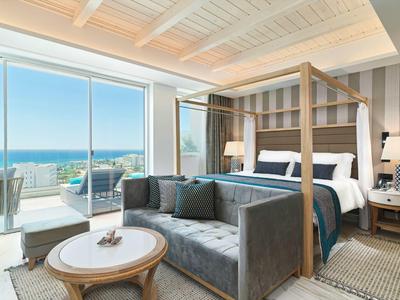 Helles Schlafzimmer mit vierpfostenbett, grauem Sofa, runder Holztisch und Meerblick durch große Fenster.