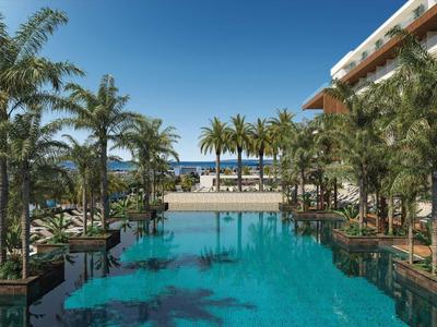 Large hotel pool with palm trees and lounge chairs under clear blue sky.