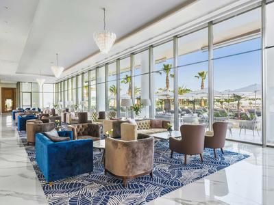 Bright lounge area with comfortable armchairs and large windows overlooking palm trees and mountains.