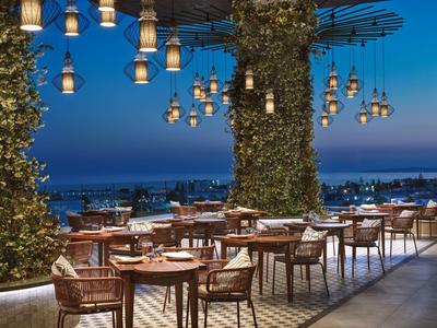 Outdoor terrace with tables, chairs, and lanterns lit at dusk overlooking the sea.