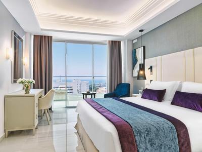 Modernes Schlafzimmer mit großem Bett, violetten Akzenten, Schreibtisch und Meerblick durch bodentiefe Fenster.