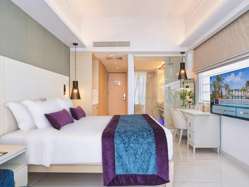 Modernes Hotelzimmer mit großem Bett, lila-blauem Überwurf und Fernseher an der Wand.