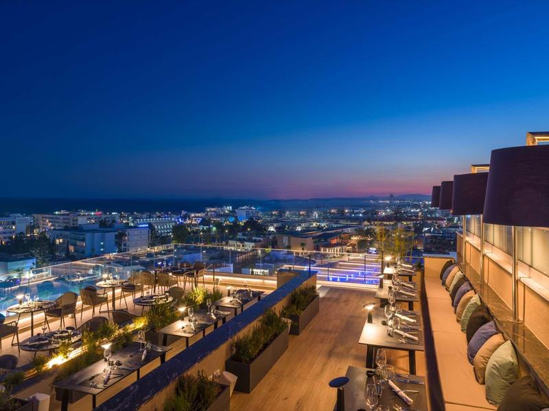 Stadtblick bei Dämmerung mit beleuchteten Straßen, Gebäuden und einer Terrasse mit Sitzgelegenheiten.