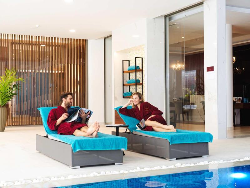 Zwei Personen entspannen auf blauen Liegen neben einem Innenpool in modernem Raum mit Holzwand.