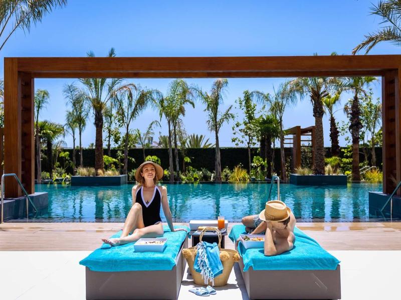 Zwei Personen entspannen auf blauen Liegen am Pool mit Palmen und blauem Himmel im Hintergrund.