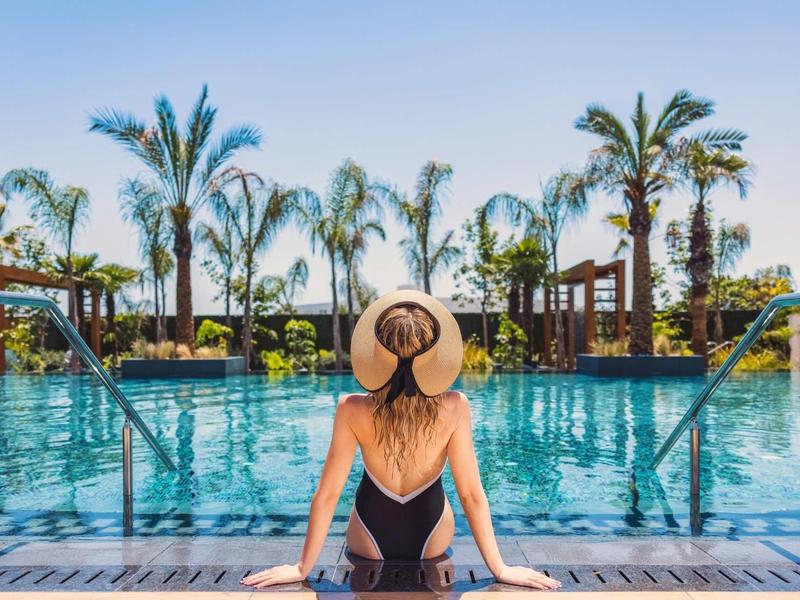 Frau mit Sonnenhut sitzt am Poolrand, umgeben von hohen Palmen und strahlend blauem Himmel.