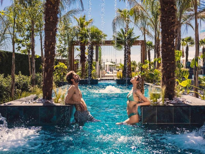 Zwei Frauen sitzen in einem blauen Außenpool, umgeben von Palmen und Wasserfontänen.