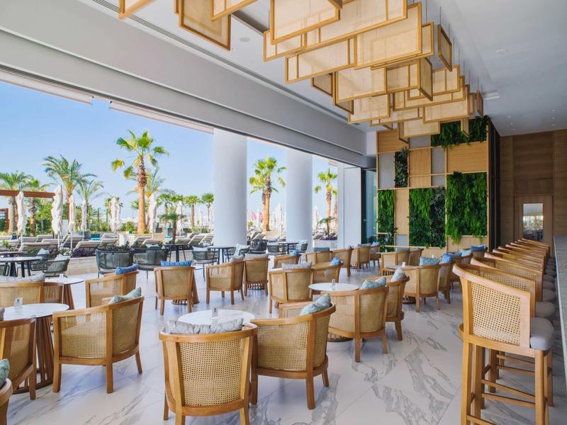 Helles Restaurant mit Holzstühlen, großen Fenstern, grünen Pflanzen und modernen Deckenlampen.