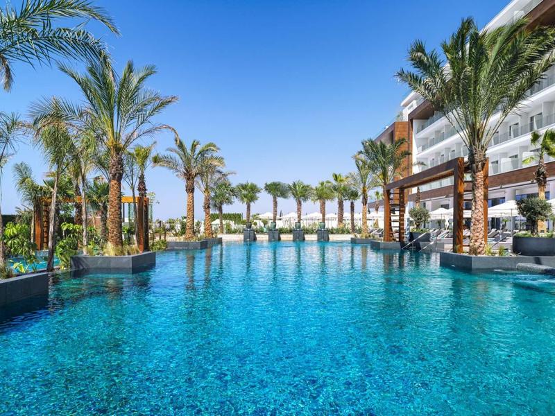 Großer blauer Pool umgeben von Palmen und modernem Hotel unter klarem, blauem Himmel.