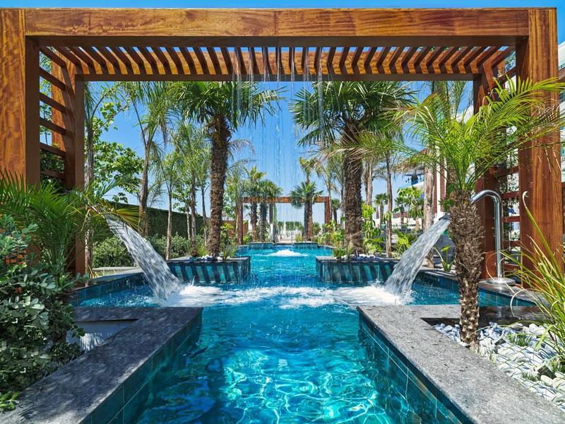 Blauer Pool mit Wasserfällen, umgeben von Palmen und Holzpergola bei sonnigem Himmel.