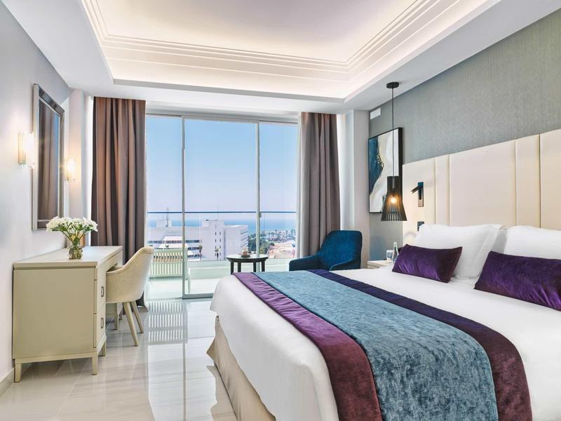 Modernes Schlafzimmer mit großem Bett, violetten Akzenten, Schreibtisch und Meerblick durch bodentiefe Fenster.