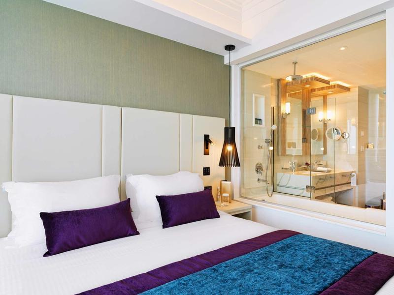 Modernes Schlafzimmer mit weißem Bett, lila Dekokissen und Glaswand zum hellen Badezimmer.