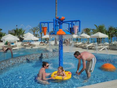 Kinder spielen in einem flachen Pool mit bunten Wasserfontänen und Sonnenschirmen im Hintergrund.