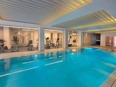 Hallenbad mit klarem blauen Wasser, moderner Decke und Fitnessgeräten hinter Glaswand.