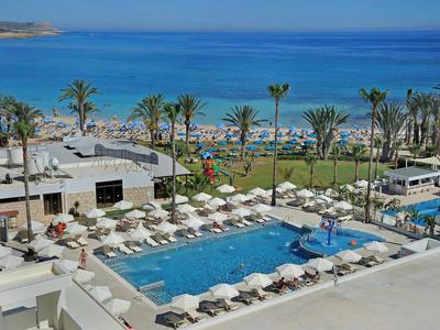Blick auf einen großen Pool mit weißen Sonnenschirmen, Palmen und blauem Meer im Hintergrund.
