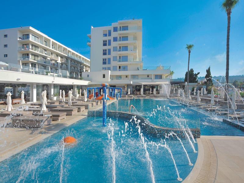 Hotel s velkým bazénem, vodními tryskami a slunečními deštníky pod modrou oblohou.