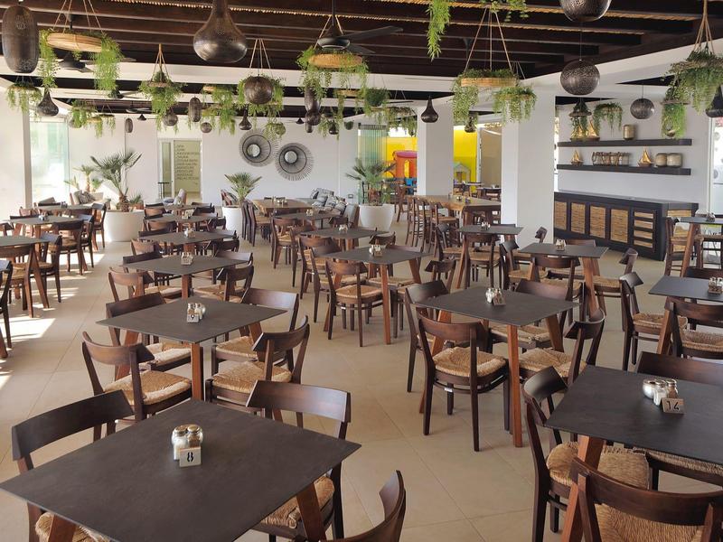 Světlá moderní hotelová restaurace s dřevěnými stoly, židlemi a zelenými rostlinami visícími ze stropu.