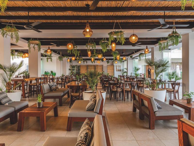 Světle zařízená hotelová jídelna s dřevěným trámovým stropem a mnoha rostlinami.