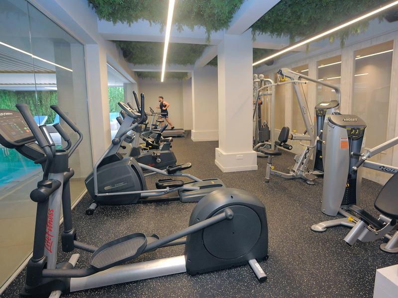 Moderní posilovna s různými cvičebními stroji a velkými okny vedle bazénu.