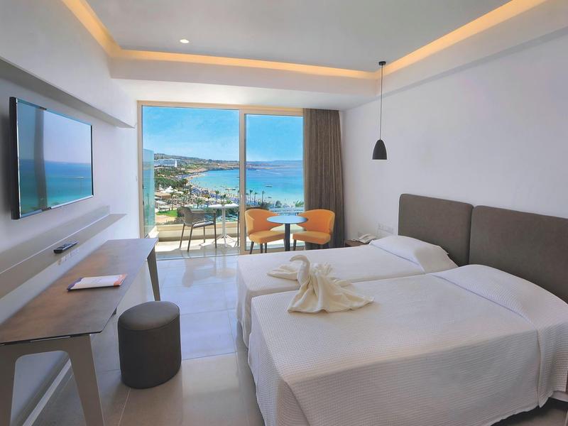 Moderní hotelový pokoj s balkonem a výhledem na moře, dvě postele a televizí na zdi.