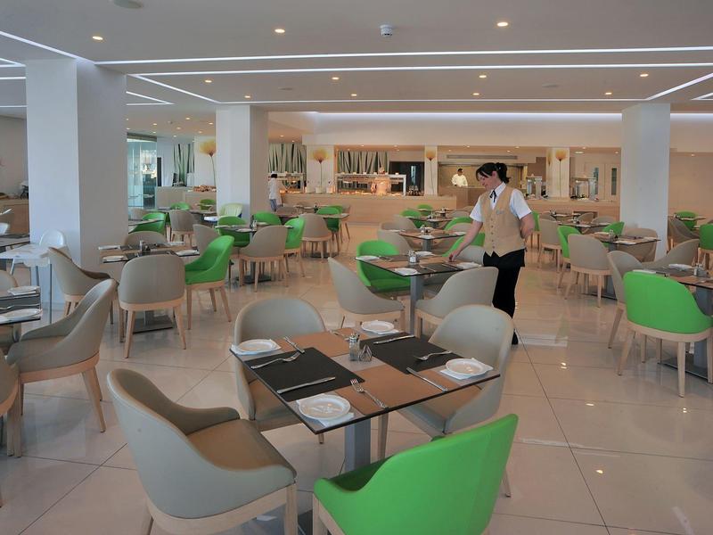 Světle zařízená moderní jídelna se zelenými a šedými židlemi a číšníkem.