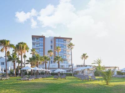 Complesso alberghiero moderno con palme e lettini su un prato verde sotto un cielo limpido.