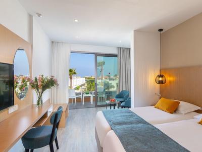 Helles Hotelzimmer mit Doppelbett, Holzmöbeln, Balkon und Blick auf das Meer.