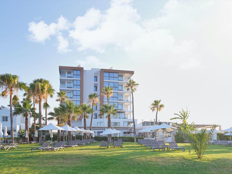 Moderner Hotelkomplex mit Palmen, Sonnenschirmen und grüner Rasenfläche unter blauem Himmel.