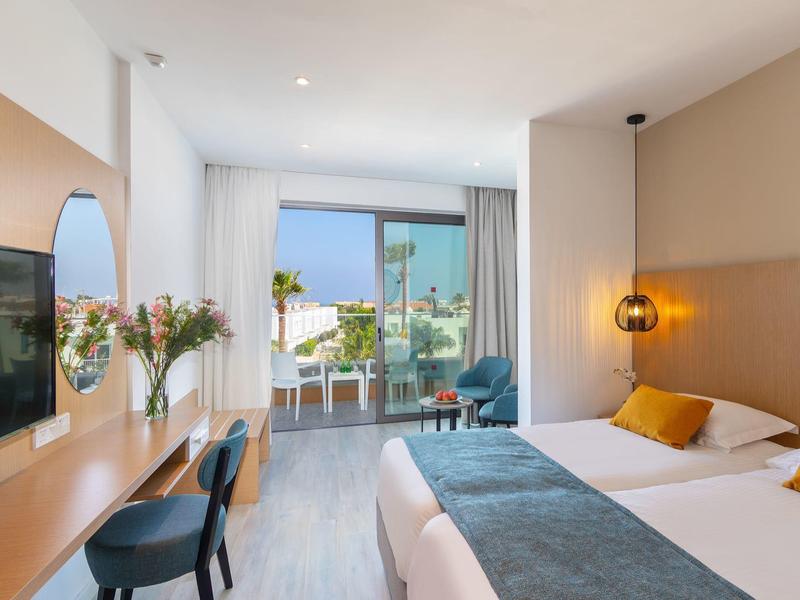 Helles Hotelzimmer mit Doppelbett, Holzmöbeln, Balkon und Blick auf das Meer.