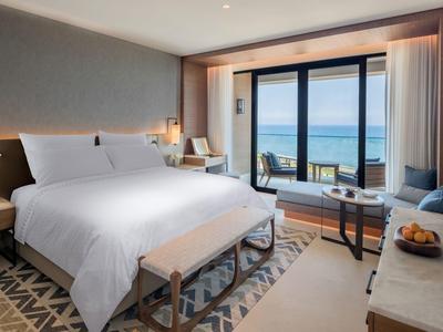 Helles Hotelzimmer mit großem Bett, Teppich, Sitzbereich und Meerblick durch Panoramascheibe.