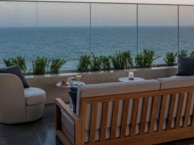 Moderne Terrasse mit Holzbett, gemütlichem Sessel, Glasgeländer und Meerblick bei Abenddämmerung.