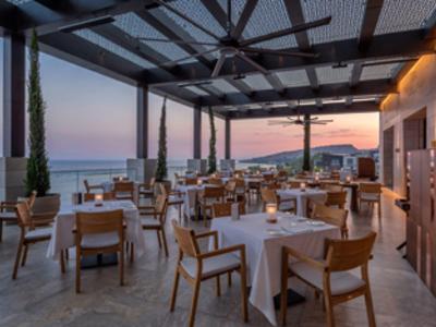 Offenes Restaurant mit Holzstühlen und weißen Tischtüchern, Meerblick bei Sonnenuntergang.