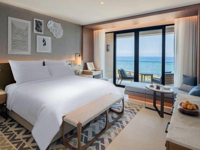 Modernes Hotelzimmer mit großem Bett, Balkon und Meerblick, helles Interieur, Teppichboden und Sitzbereich.