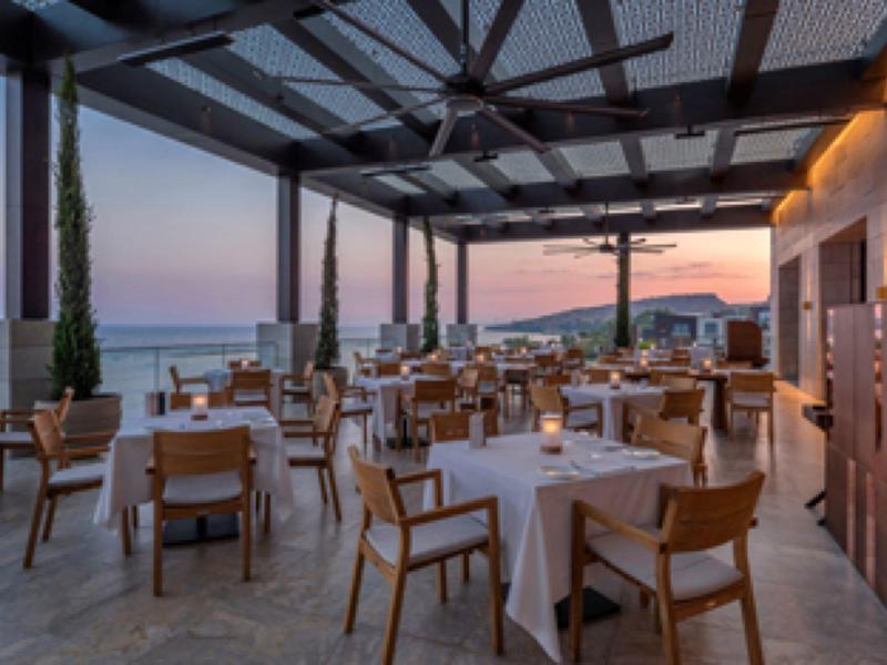 Offenes Restaurant mit Holzstühlen und weißen Tischtüchern, Meerblick bei Sonnenuntergang.