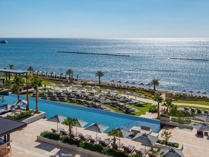 Blick auf einen Pool, Liegestühle und Palmen am Strand mit ruhigem, blauem Meer und klarem Himmel.