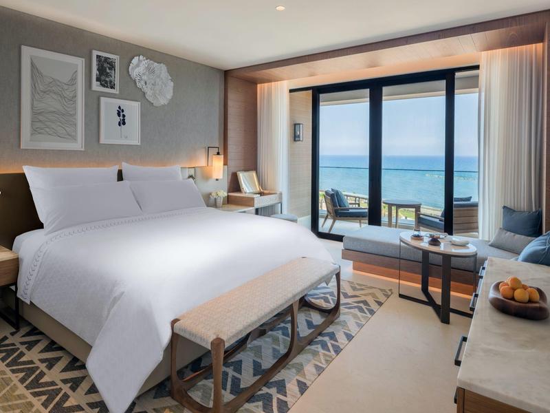 Modernes Hotelzimmer mit großem Bett, Balkon und Meerblick, helles Interieur, Teppichboden und Sitzbereich.