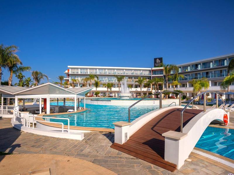 Hotelový komplex s velkým bazénem, mostkem, lehátky a palmami pod jasnou modrou oblohou.