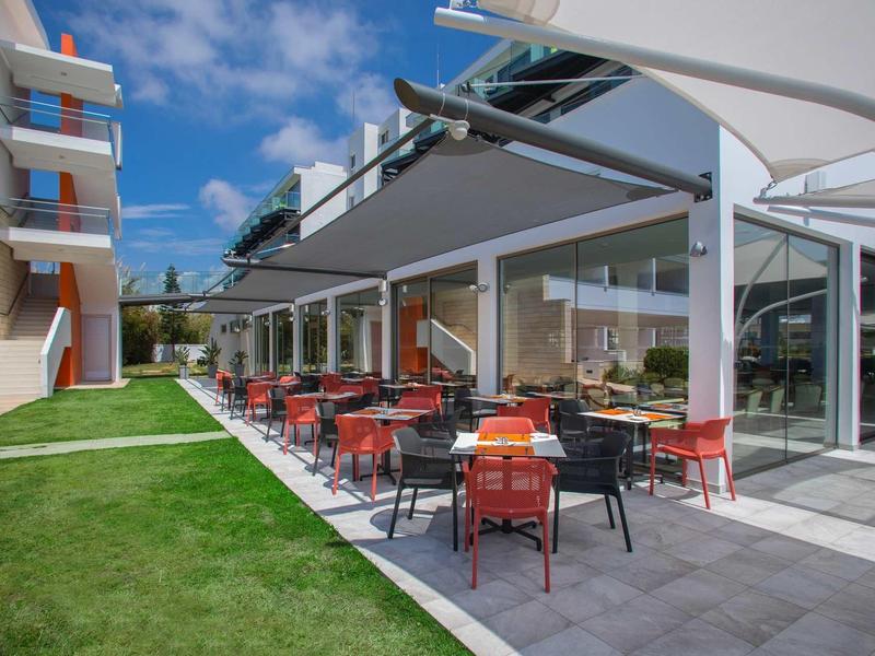 Moderní venkovní prostor hotelu s stoly, židlemi a zeleným trávníkem pod modrou oblohou.