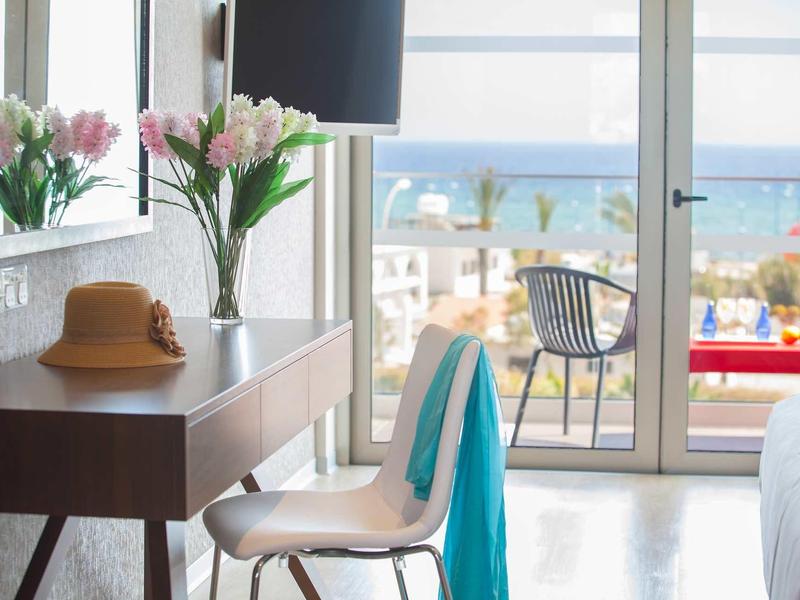Světý hotelový pokoj s výhledem na moře, psacím stolem, židlí a květinami na komodě.