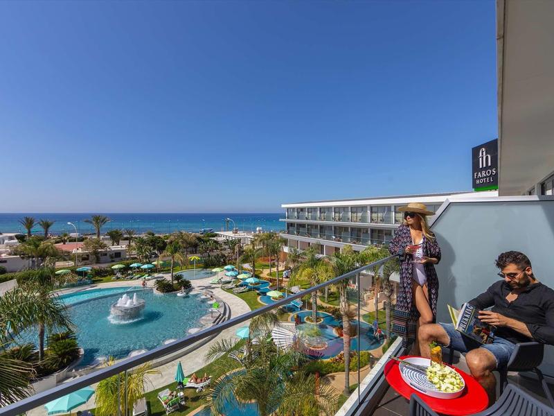 Pohled z hotelového balkonu na bazén, zahradu a moře za slunečného dne.