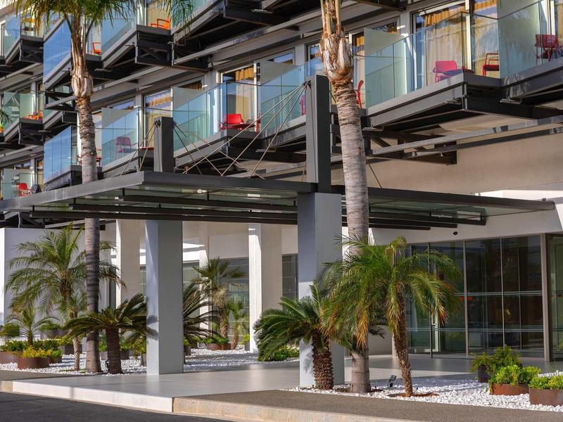 Vnější pohled na moderní hotel s balkony a palmami u vchodu.