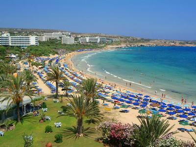 Ein langer Sandstrand mit vielen blauen Sonnenschirmen und Palmen neben einem Hotel.