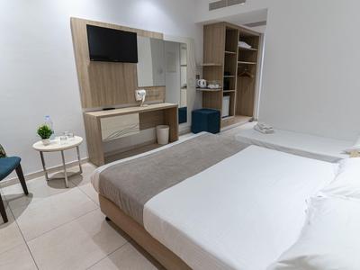 Modern hotel room with double bed, TV, desk, and open wardrobe in neutral tones.